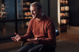 Smaller image of man in orange sweatshirt sitting down and speaking right to off-camera